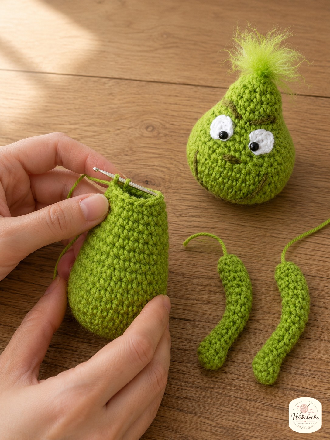 Länglicher grüner Körper und dünne Arme entstehen neben dem fertigen Kopf des Grinch Amigurumi